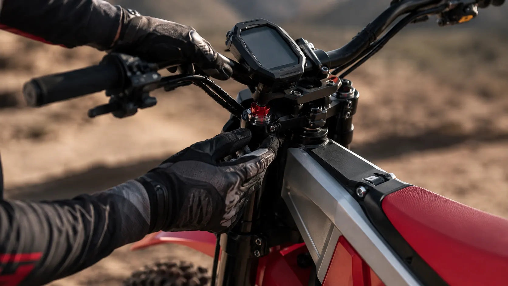 Close-up of gloved hands performing technical adjustments on the central triple clamp or suspension forks of a NATKIE high-tech electric dirt bike on a dusty outdoor trail.