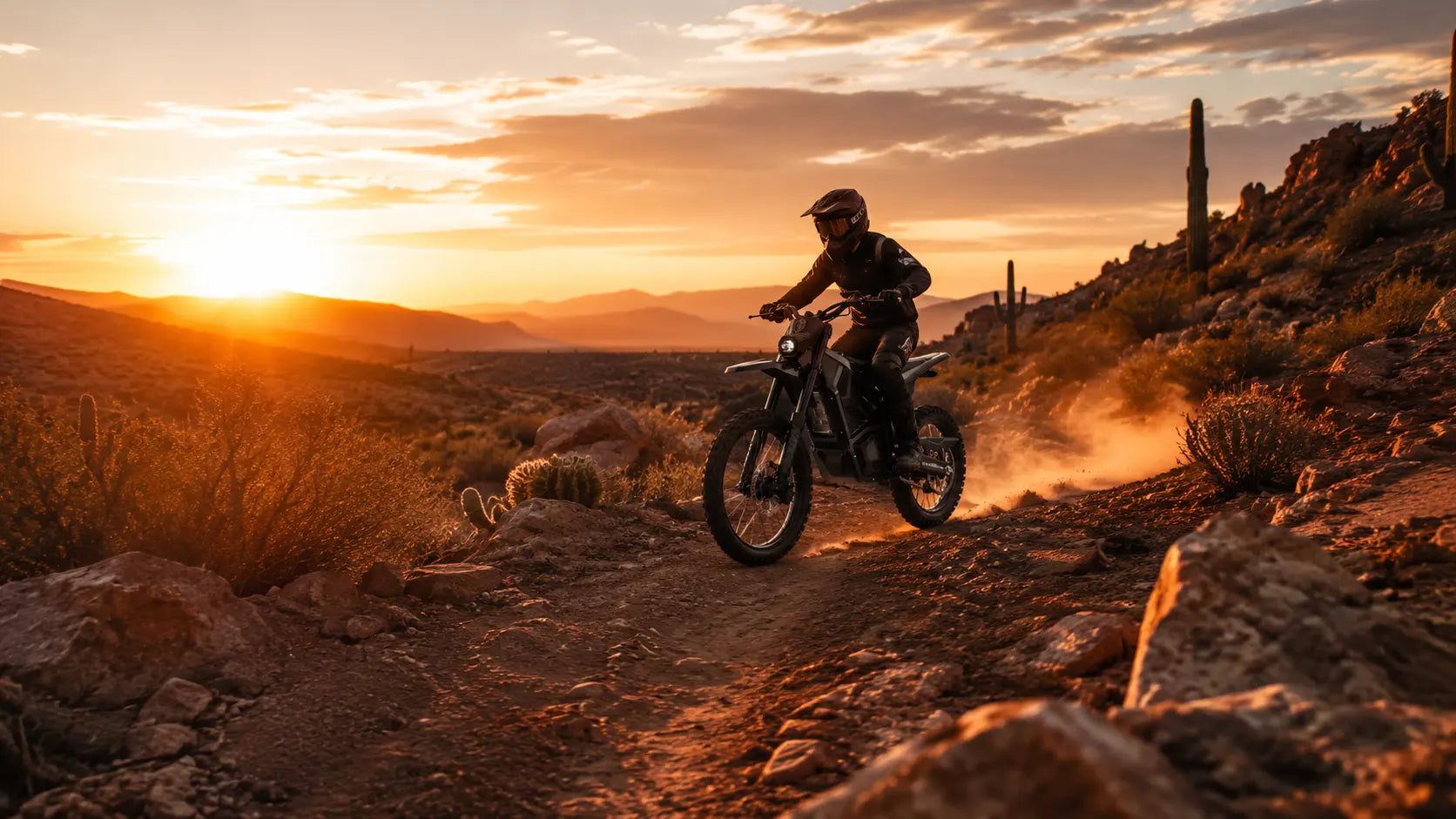 A wide landscape shot of a rugged, rugged desert OHV trail in the Sonoran Desert at sunset, where a rider on a sleek black electric dirt bike is kicking up a small trail of dust. Saguaro cacti are visible in the distance.